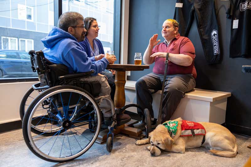Photo of three people around a cafe table. One person is in a wheelchair. One person has a guide dog at this feet and is engaging in conversation.